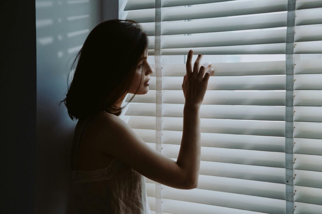 A young woman in a nightdress gazes thoughtfully through window blinds, creating a serene and introspective mood.