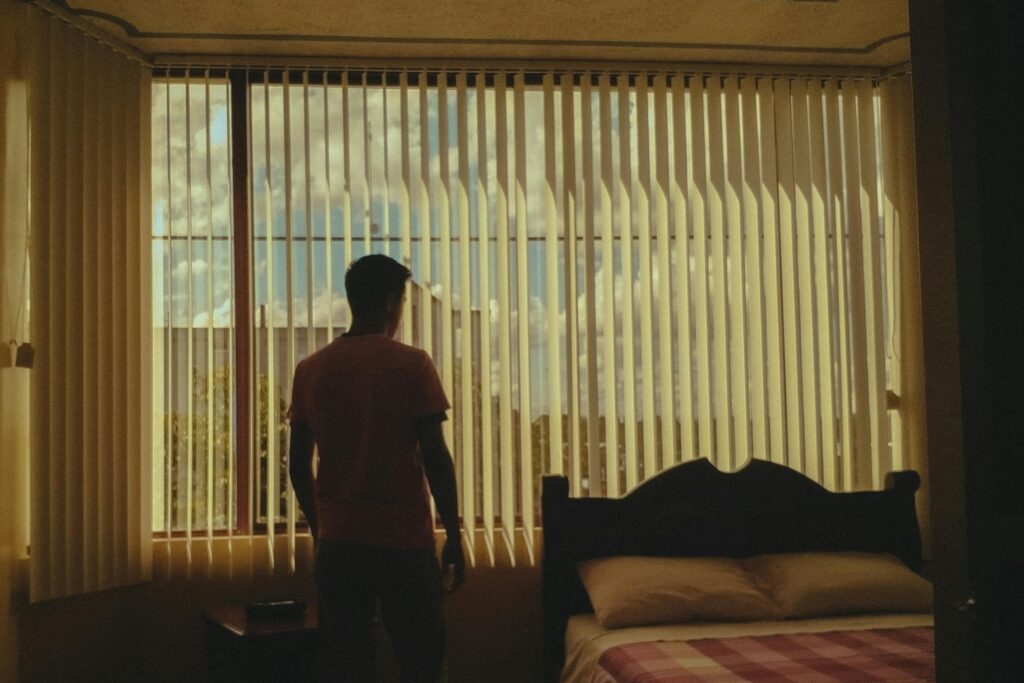 A silhouette of a man stares out a bedroom window with closed blinds, emphasizing introspection.